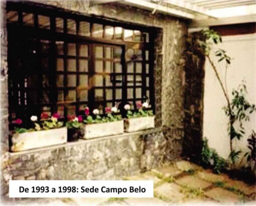 Esta imagem retrata a Sede Campo Belo entre os anos de 1993 a 1998. O edifício antigo exibe uma janela gradeada com flores coloridas no peitoril e plantas ao redor. A construção parece ser feita de pedra ou concreto áspero, conferindo-lhe uma aparência envelhecida e robusta. As flores adicionam um toque de cor à cena, enquanto as plantas ao lado da janela sugerem um ambiente natural ou um jardim próximo. A legenda “De 1993 a 1998: Sede Campo Belo” está escrita na parte inferior da imagem, indicando o período de tempo e localização específicos retratados na foto.