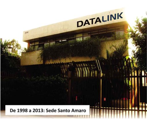 Esta imagem retrata a Sede Santo Amaro da DATALINK entre os anos de 1998 a 2013. O edifício, de dois andares, é branco e exibe a inscrição “DATALINK” no topo. Vegetação cresce nas laterais do edifício, e um portão de ferro preto está localizado na frente. O número “314” é visível na parte superior esquerda do edifício. A legenda “De 1998 a 2013: Sede Santo Amaro” está na parte inferior da imagem, indicando o período em que o edifício foi utilizado como sede da empresa. O céu ao fundo está claro, sugerindo que a foto foi tirada durante o dia.