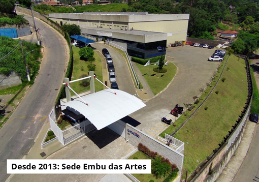 Esta imagem retrata a Sede Embu das Artes desde 2013. O complexo moderno inclui um edifício principal, estacionamento e uma entrada coberta. O edifício principal tem um design contemporâneo, com cores cinza claro e escuro. Ao lado, há um estacionamento com vários carros estacionados. Uma entrada coberta branca está localizada na frente do complexo, conectada por uma estrada pavimentada que serpenteia pelo terreno. O ambiente é bem cuidado, com gramados verdes e árvores ao redor. O céu claro sugere que a foto foi tirada durante o dia.