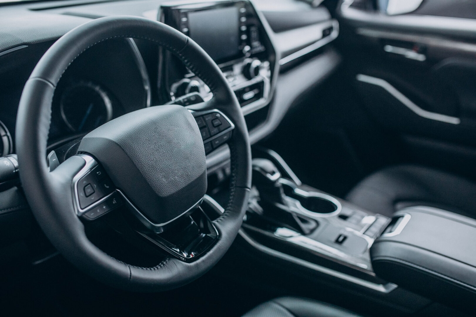 Fotografia mostrando o interior de um carro moderno em tons escuros. Em primeiro plano, vê-se um volante revestido em couro e, ao centro, o painel de controle com tela digital. Mais à direita, ficam a alavanca de câmbio e porta-copos no console central."