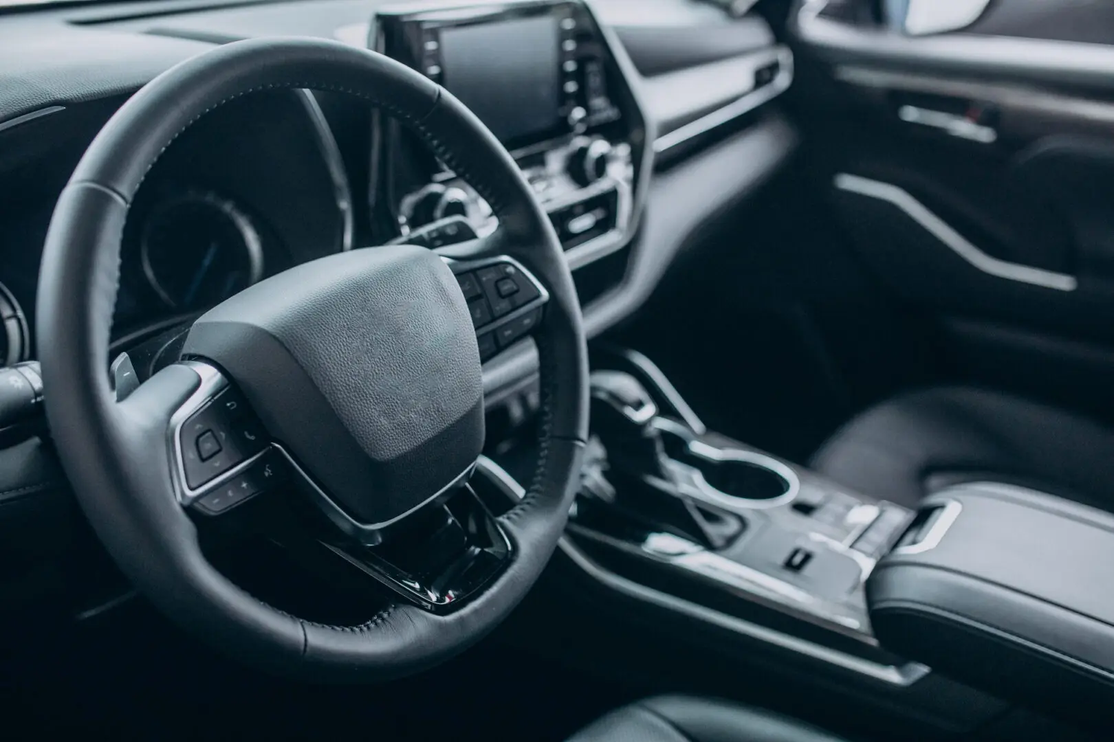 26488.jpg Fotografia mostrando o interior de um carro moderno em tons escuros. Em primeiro plano, vê-se um volante revestido em couro e, ao centro, o painel de controle com tela digital. Mais à direita, ficam a alavanca de câmbio e porta-copos no console central."
