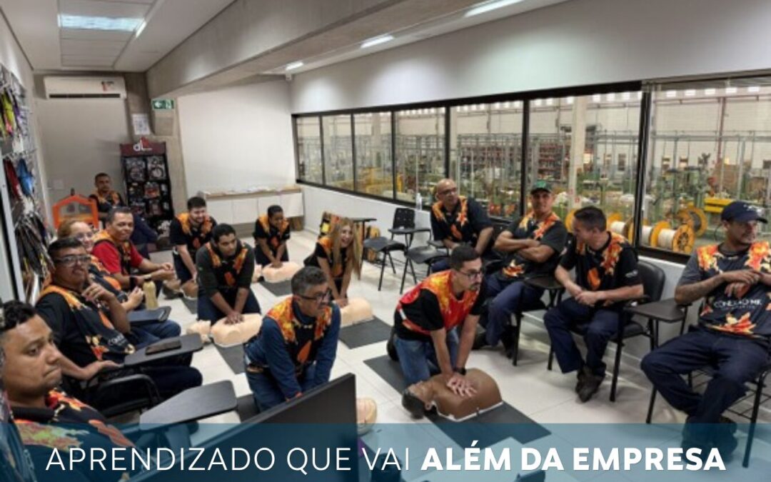 Group of employees in matching vests sit in a training room around CPR mannequins, engaged in a hands-on with an industrial workshop visible through windows in the background.