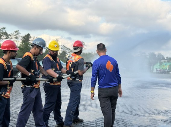 Imagem3 Five trainees in helmets hold hoses while an instructor watches as water sprays on a wet tarmac.