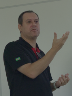 Man in a black polo with a Brazil flag patch on the sleeve, gesturing with his right hand while speaking.