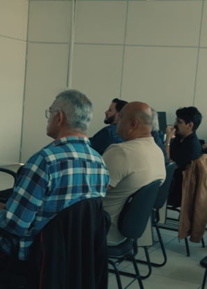Adults seated in a classroom-style room watching a presentation; man in a blue plaid shirt in foreground.